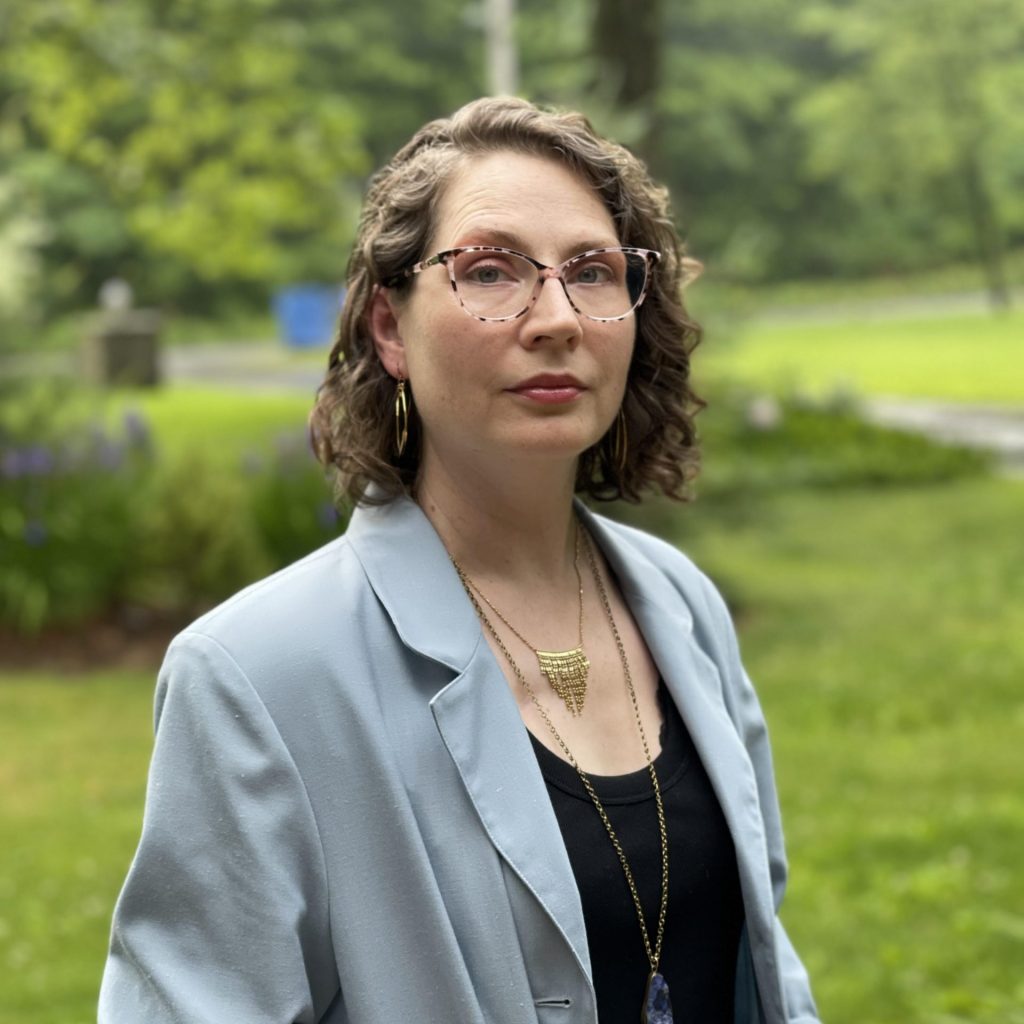 Woman with glasses and short hair poses against green grass background in headshot pose.